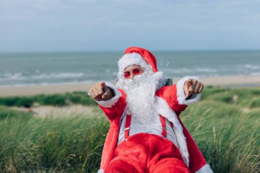 Deniz kenarındaki iskemlede oturan gülümseyen Noel Baba işaret parmağıyla seni tatile davet ediyor. Parmaklarına odaklan.