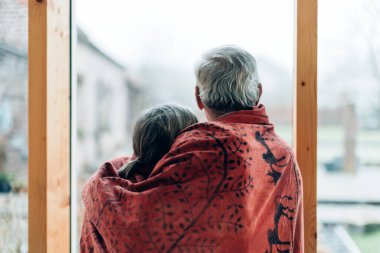 Sarılmış yaşlı bir çiftin sırtından vurularak büyük camdan bir duvardan dışarı bakan bir battaniyeye sarılmış.