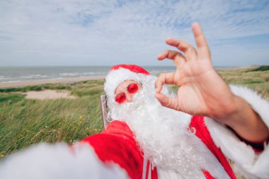 Noel Baba tatilde. Noel Baba gibi giyinmiş bir adam iskemlede oturuyor ve selfie çekiyor.