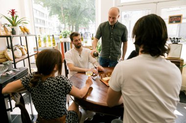 Restoran sahibi bardaklara şarap dolduruyor. Müşteriler masanın etrafında oturuyor.