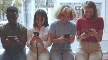 Çok ırklı gençler pencere eşiğinde yan yana otururken cep telefonu kullanıyorlar. Teknoloji kullanarak yakınlaştırıyorlar.