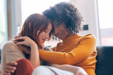 Arka planda oturan lezbiyen çift kanepede sarılıyor.