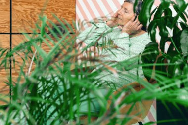 Yaşlı kadın, yeşil bitkilerle çevrili kulaklıklarıyla rahat bir sandalyede oturmuş müzik dinliyor.