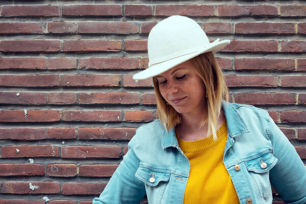 caucasian blond woman wearing white hat - denim jacket - yellow sweater against a brick wall background