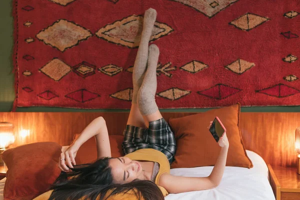 asian woman in miniskirt and thigh-high socks lying on the bed with legs up against the wall holding and looking at mobile phone