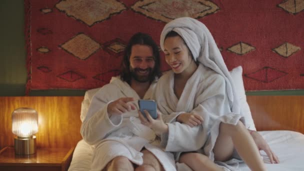 多民族夫妇坐在床上 穿着浴衣 笑着看手机 — 图库视频影像