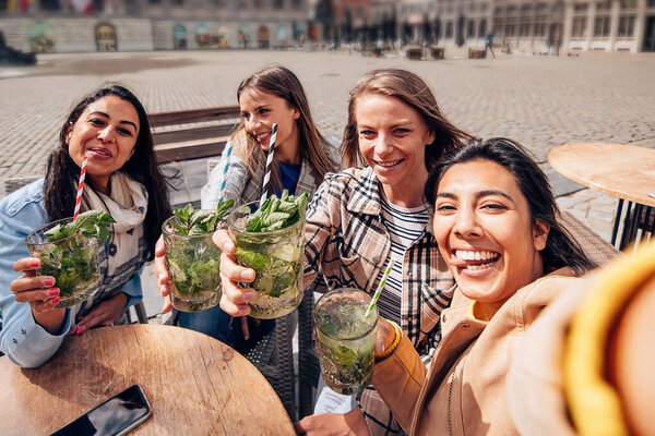 group of multiethnic young happy cheerful women sitting on terrace of the city center with cocktails in their hands taking a selfie