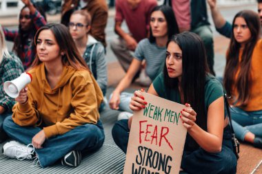 Sokakta oturan genç kalabalık cinsiyet eşitliğini protesto ediyor. Zayıf erkeklerle dolu panel güçlü kadınlardan korkuyor.