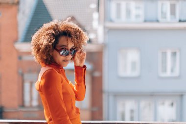 Şehrin balkonunda güneş gözlüklü modaya uygun kıvırcık kız.