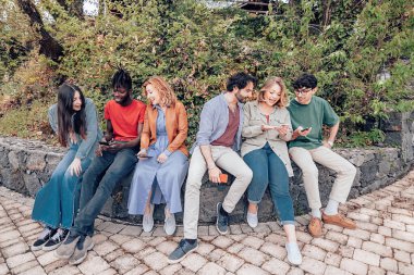 Alçak bir duvarda oturan, konuşan ve cep telefonlarına bakan bir grup çok ırklı arkadaş.