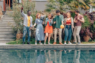 Bir grup arkadaş gündüz partisinde müzik, bira ve klasik müzik setiyle havuz başında dans ediyor.
