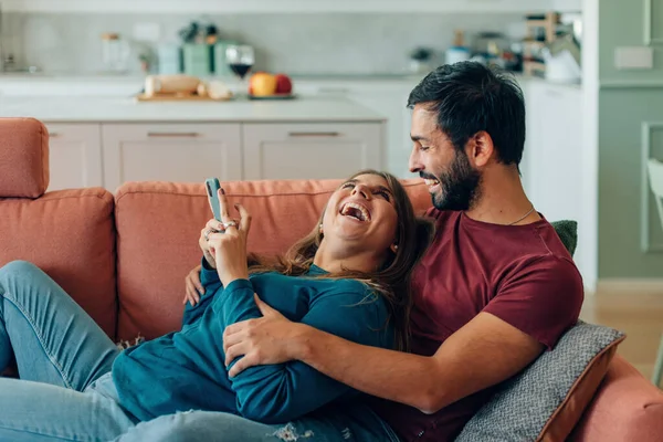 Lykkeligt Par Kramme Ved Hjælp Mobiltelefon Sammen Liggende Sofaen Derhjemme - Stock-foto