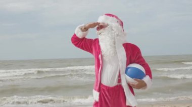 Noel Baba deniz kıyısında tatilde plaj topuyla sahilde dikiliyor.