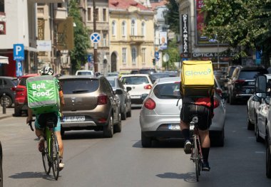 Bucharest, Romania - July 04, 2022: Bolt Food and Glovo food delivery couriers ride bicycles on a street through car traffic.