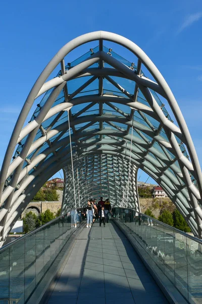 Tbilisi, Geórgia - 24 de outubro de 2021: Ponte da Paz, ponte pedonal em forma de arco sobre o rio Kura, na capital georgiana. Construção de aço e vidro. Pessoas caminhando na ponte — Fotografia de Stock