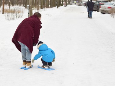 Anne ve küçük oğlu parkta çocukların plastik kayaklarında kayak yapıyorlar. Parkta kış tatili.