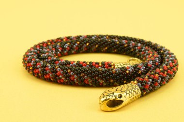 Yılan başı ve kuyruğu şeklinde uçlarında metal tokalar bulunan çok renkli boncuklardan örülmüş kadın mücevherleri. Sarı arkaplanda izole edilmiş.