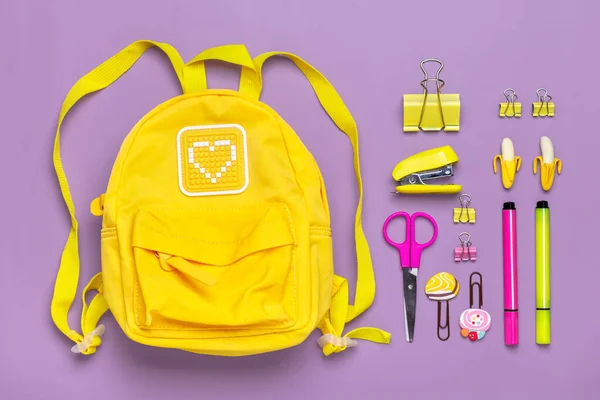 Wracając do szkoły, koncepcja edukacji. Żółty plecak z materiałami szkolnymi - notatnik, długopisy, klipsy, nożyczki izolowane na fioletowym tle — Zdjęcie stockowe