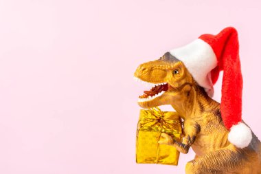 Kırmızı Noel Baba şapkalı dinozor Rex 'in pençelerinde altın hediye kutusu var. Yeni yıl arifesinde ya da Noel arifesinde. Mutlu Noeller için yaratıcı bir fikir..