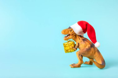 Kırmızı Noel Baba şapkalı dinozor Rex mavi arka planda altın hediye kutusu tutuyor. Yeni yıl arifesi ya da Noel arifesi resmi. Mutlu Noel konsepti için yaratıcı bir fikir..