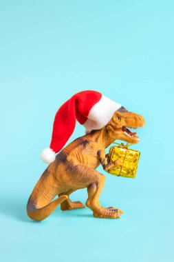 Kırmızı Noel Baba şapkalı dinozor Rex mavi arka planda altın hediye kutusu tutuyor. Yeni yıl arifesi ya da Noel arifesi resmi. Mutlu Noel konsepti için yaratıcı bir fikir..