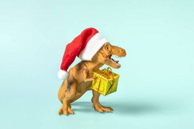 Kırmızı Noel Baba şapkalı dinozor Rex mavi arka planda altın hediye kutusu tutuyor. Yeni yıl arifesi ya da Noel arifesi resmi. Mutlu Noel konsepti için yaratıcı bir fikir..