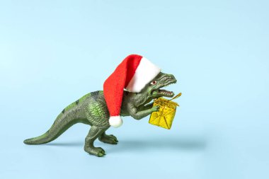 Kırmızı Noel Baba şapkalı dinozor Rex mavi arka planda altın hediye kutusu tutuyor. Yeni yıl arifesi ya da Noel arifesi resmi. Mutlu Noel konsepti için yaratıcı bir fikir..