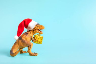 Kırmızı Noel Baba şapkalı dinozor Rex mavi arka planda altın hediye kutusu tutuyor. Yeni yıl arifesi ya da Noel arifesi resmi. Mutlu Noel konsepti için yaratıcı bir fikir..