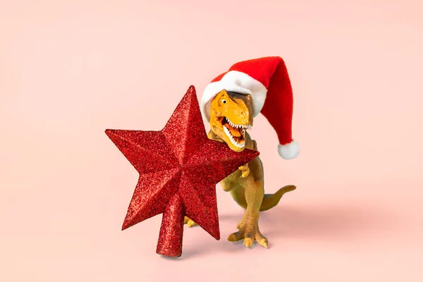 赤いサンタクロースの帽子の恐竜レックスピンクの背景にその爪で星を保持大晦日やクリスマスイブアートホリデーカードメリークリスマスのコンセプトのための創造的なアイデア — ストック写真