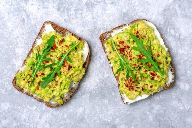 Kahvaltı için sandviç hazırla - bir dilim kepekli ekmek, krem peynir, guakomole, roka ile süslenmiş, beton masa üzerinde kırmızı biber düz konsept ev yapımı gıda konsepti