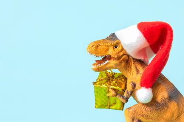Kırmızı Noel Baba şapkalı dinozor Rex mavi arka planda altın hediye kutusu tutuyor. Yeni yıl arifesi ya da Noel arifesi resmi. Mutlu Noel konsepti için yaratıcı bir fikir..