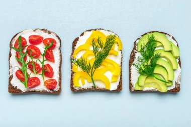 Kahvaltı için çeşitli sandviçler - bir dilim kepekli ekmek, sarı dolma biber, krem peynir, avokado, kiraz domatesleri, dereotla süslenmiş, yeşil roka mavi masanın üstünde..