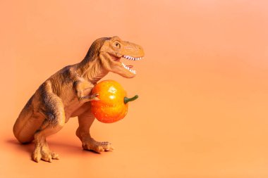 Toy dinosaur Tyrannosaurus holding pumpkin in its paws isolated on orange background Holiday greeting card Happy Halloween day, Hello Autumn creative minimal concept.
