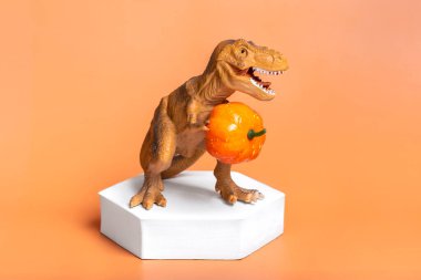 Toy dinosaur Tyrannosaurus holding pumpkin in its paws, on white podium isolated on orange background Holiday greeting card Happy Halloween day, Hello Autumn creative minimal concept.