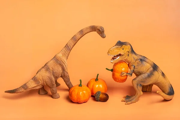 Toy dinosaur Tyrannosaurus holding pumpkin in its paws, on white podium isolated on orange background Holiday greeting card Happy Halloween day, Hello Autumn creative minimal concept.