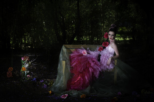 Beautiful fantasy woman with flowers and pearls in her dark hair, reclining on wooden bench in forest