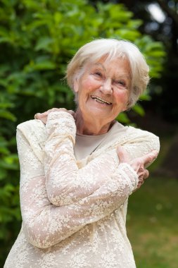 Elderly woman hugging herself with her arms in a garden clipart