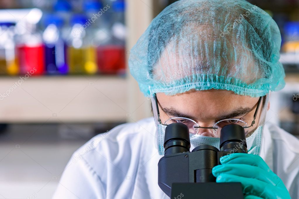 Male chemist working in a laboratory wearing glasses, a surgical cap and mask looking down the ocular of his microscope as he performs an analysis   close up of his head.
