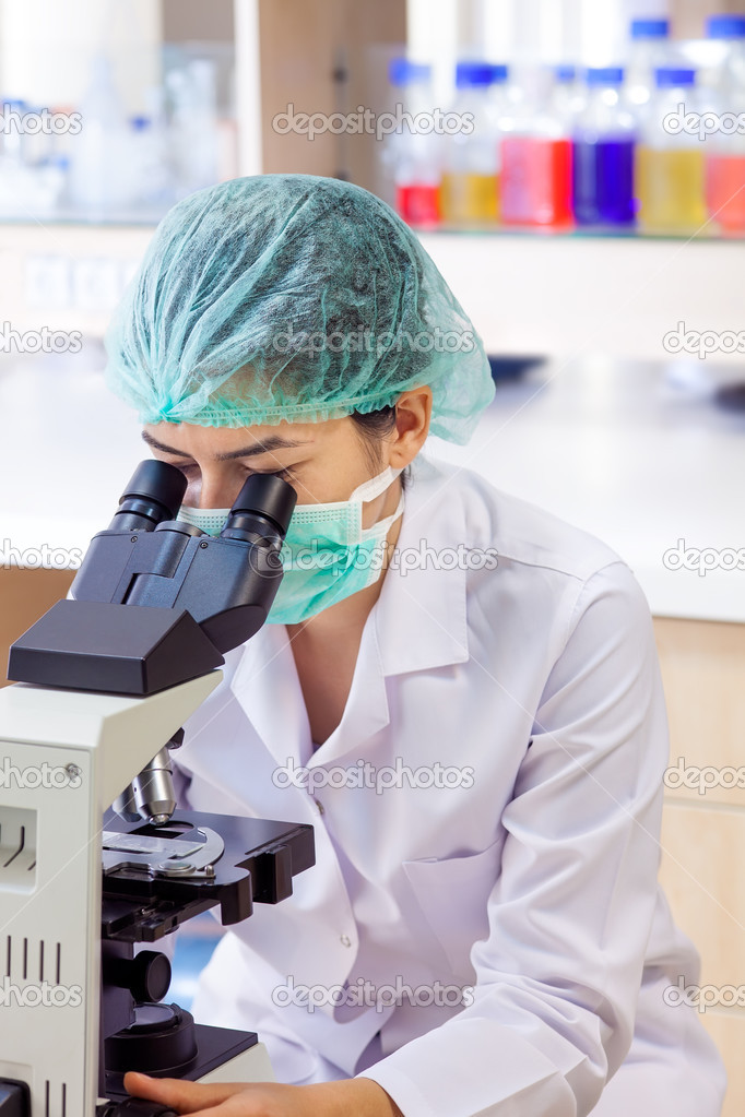 Laboratory technologist or technician using a microscope, analysing a slide while wearing a surgical cap and mask.