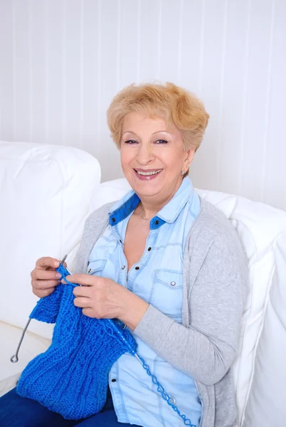 Счастливая улыбающаяся пожилая женщина вяжет на диване дома — стоковое фото