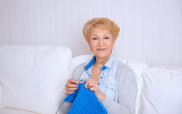 Felice donna anziana sorridente che lavora a maglia sul divano a casa — Foto Stock