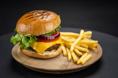Tasty homemade beef burger with fresh ingredients served on little wooden cutting board with french fries on dark background, close-up view.	