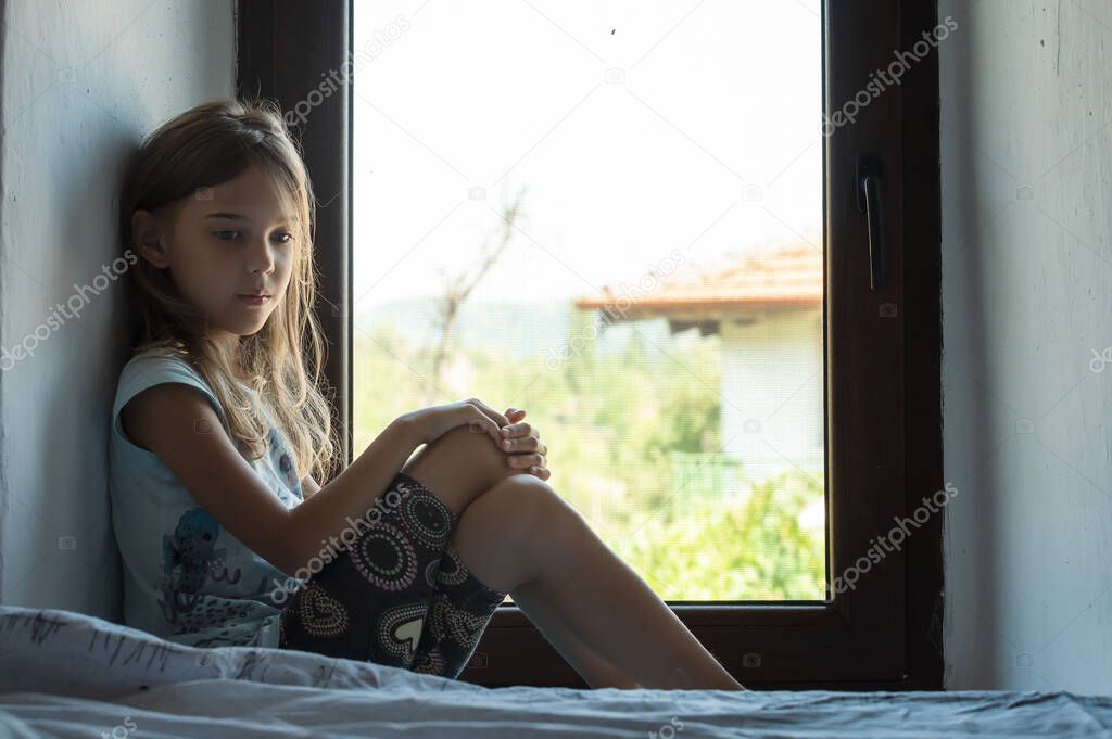 Vintage portrait of a beautiful little girl with bare feet and pensive gaze sitting near a window inside a room