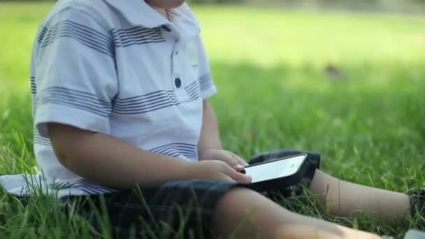 Jovem menino bonito usando telefone tela sensível ao toque no parque . — Vídeo de Stock