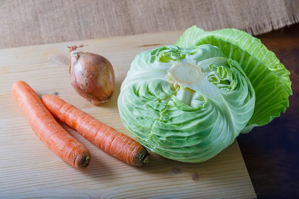 Las zanahorias crudas, la cebolla con la cáscara y la col blanca con las hojas verdes sobre la tabla de madera. Comida vegetariana saludable. —  Fotos de Stock
