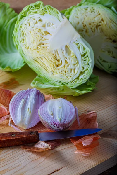 Verduras frescas crudas cortadas en dos partes. Cebolla morada con cáscara y col blanca en una tabla de madera, un cuchillo pequeño. —  Fotos de Stock