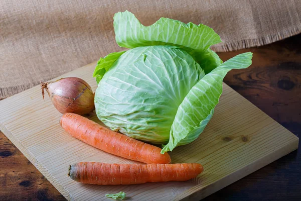 Verduras crudas zanahorias, una cebolla con cáscara y col blanca con hojas verdes en una tabla de madera. Comida vegetariana saludable. — Foto de Stock