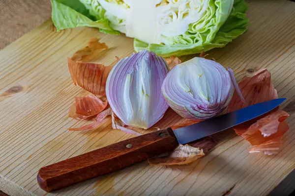 Vista superior de verduras crudas cortadas en dos partes. Cebolla morada con cáscara y col blanca en una tabla de madera, un cuchillo pequeño. —  Fotos de Stock