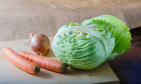Hortalizas crudas la col con las hojas verdes, la zanahoria, la cebolla sobre la tabla de madera. Un paño de arpillera. Comida vegetariana saludable. — Foto de Stock