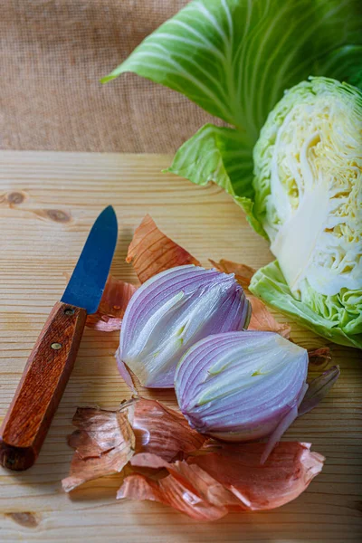 Cebolla morada con cáscara y col blanca en una tabla de madera, un cuchillo pequeño. Verduras frescas crudas cortadas en dos partes. —  Fotos de Stock